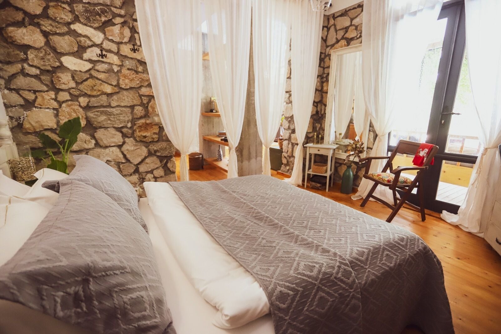 A bedroom with a stone wall, a bed with patterned bedding, a small table with a vase, and a chair by a window with curtains.