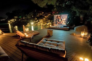 A wooden deck with cushioned seating, a screen displaying a couple, and ambient lighting. - Nautical Hotel Adults Only  (Fethiye)