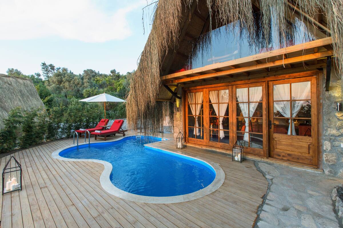 Una terraza de madera con piscina, sillas reclinables rojas y un edificio con techo de paja.