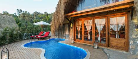 Eine Holzterrasse mit Swimmingpool, roten Liegestühlen und einem Gebäude mit Strohdach.