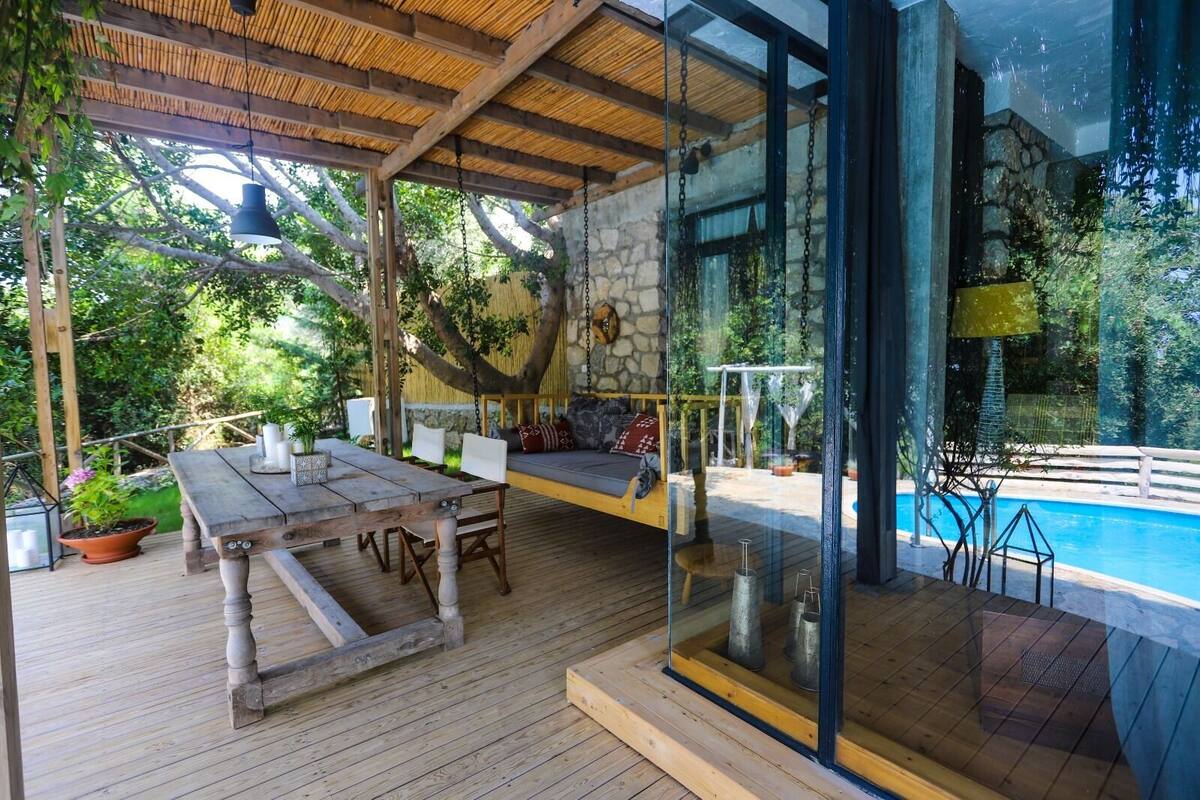 Una terraza de madera con una mesa y sillas, una mecedora colgante y un muro de cristal con vistas a una piscina.