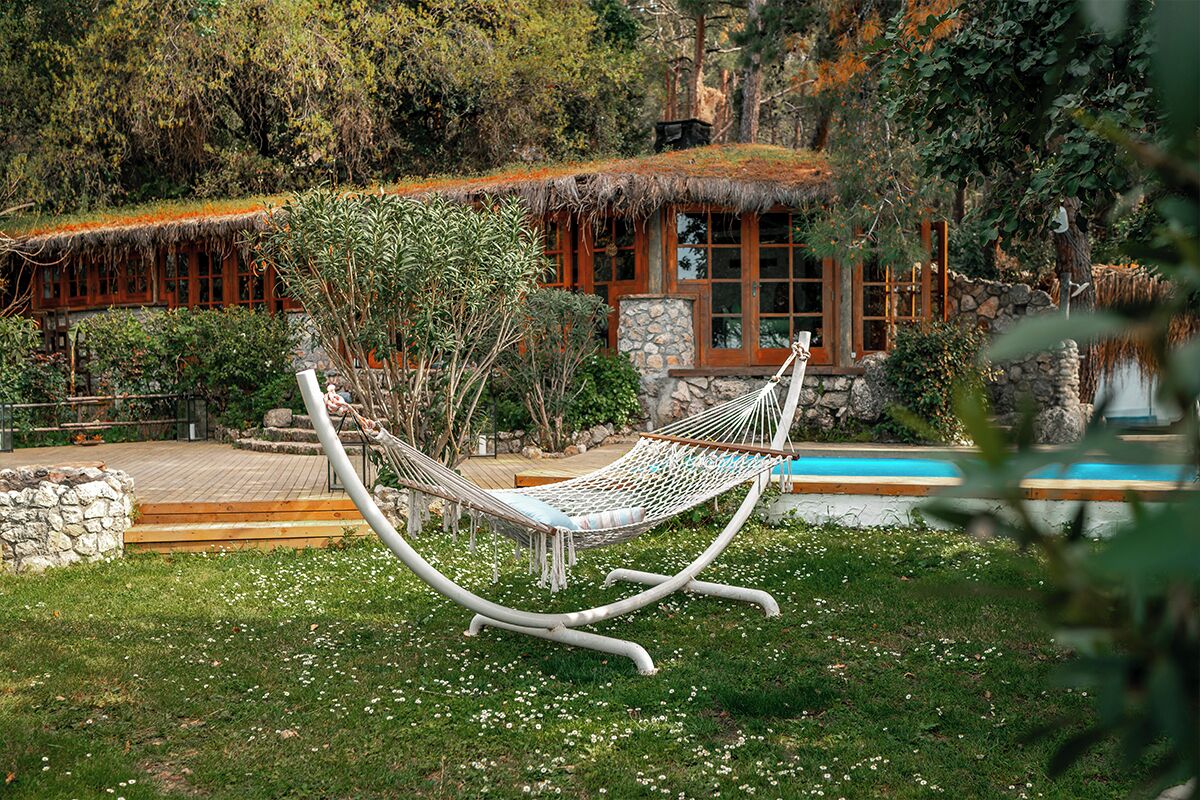 A hammock in a lush garden with a stone wall and a pool in the background.