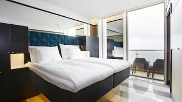 Una habitación de hotel moderna con una cama grande, un cabecero oscuro y vistas al balcón a través de un ventanal.