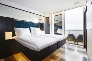 Una habitación de hotel moderna con una cama grande, un cabecero oscuro y vistas al balcón a través de un ventanal.