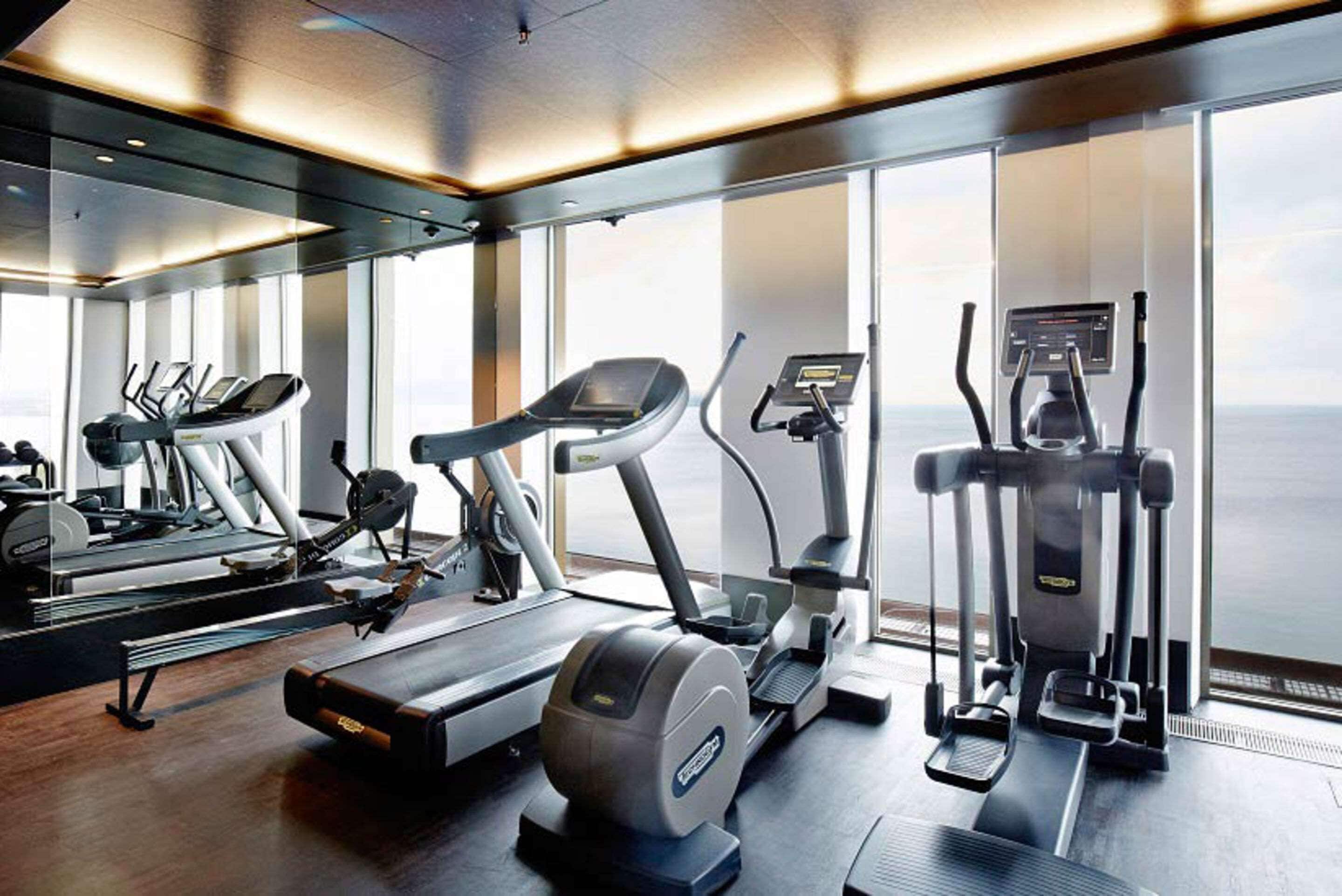 A modern gym with various cardio machines, large windows, and a wooden floor.