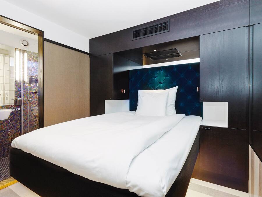 Una habitación de hotel moderna con una cama grande, un escritorio integrado y un baño con un vibrante mosaico en el fondo.