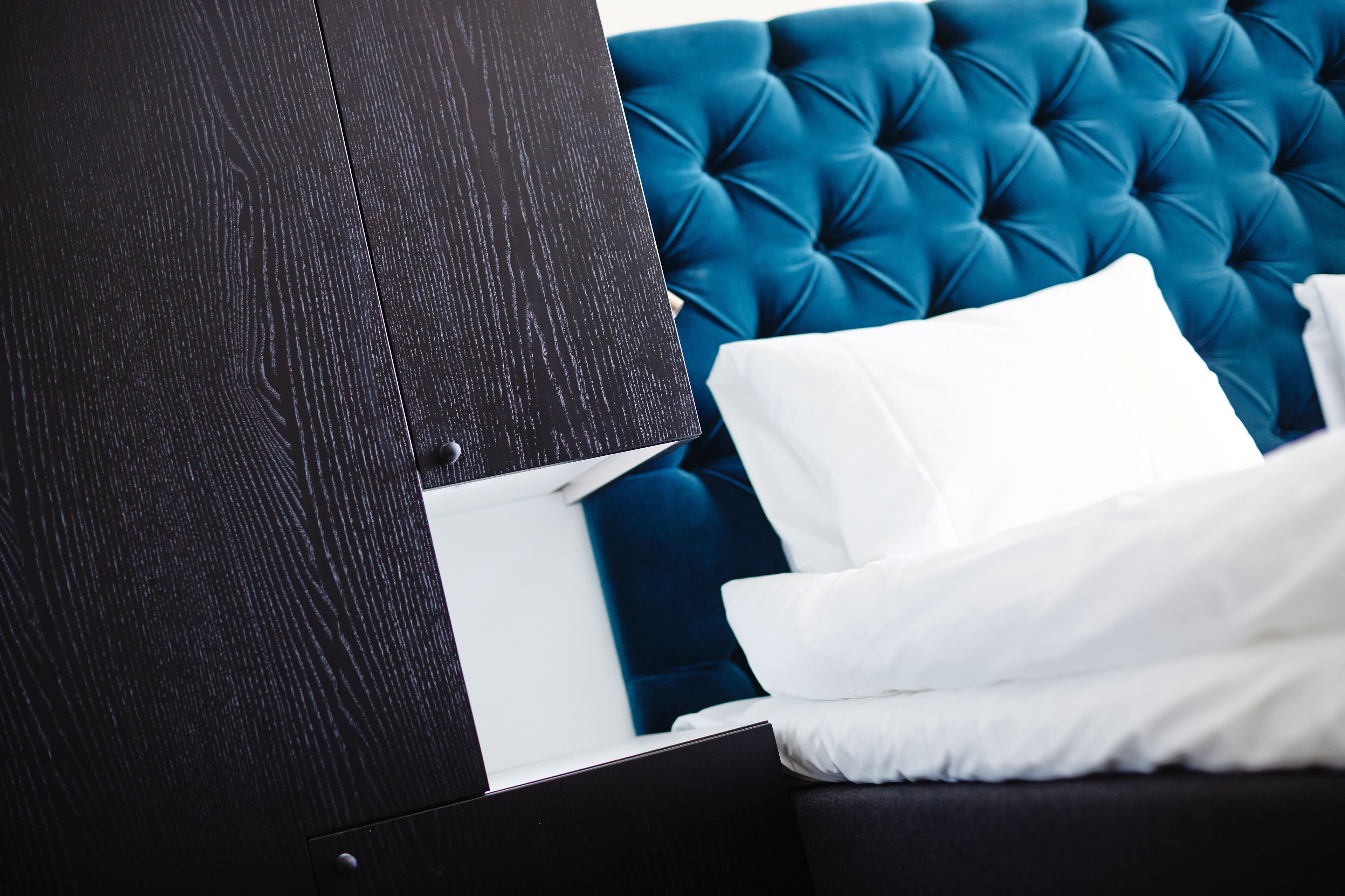 Una habitación moderna con un armario de madera oscura, un cabecero azul con capitonado y ropa de cama blanca.