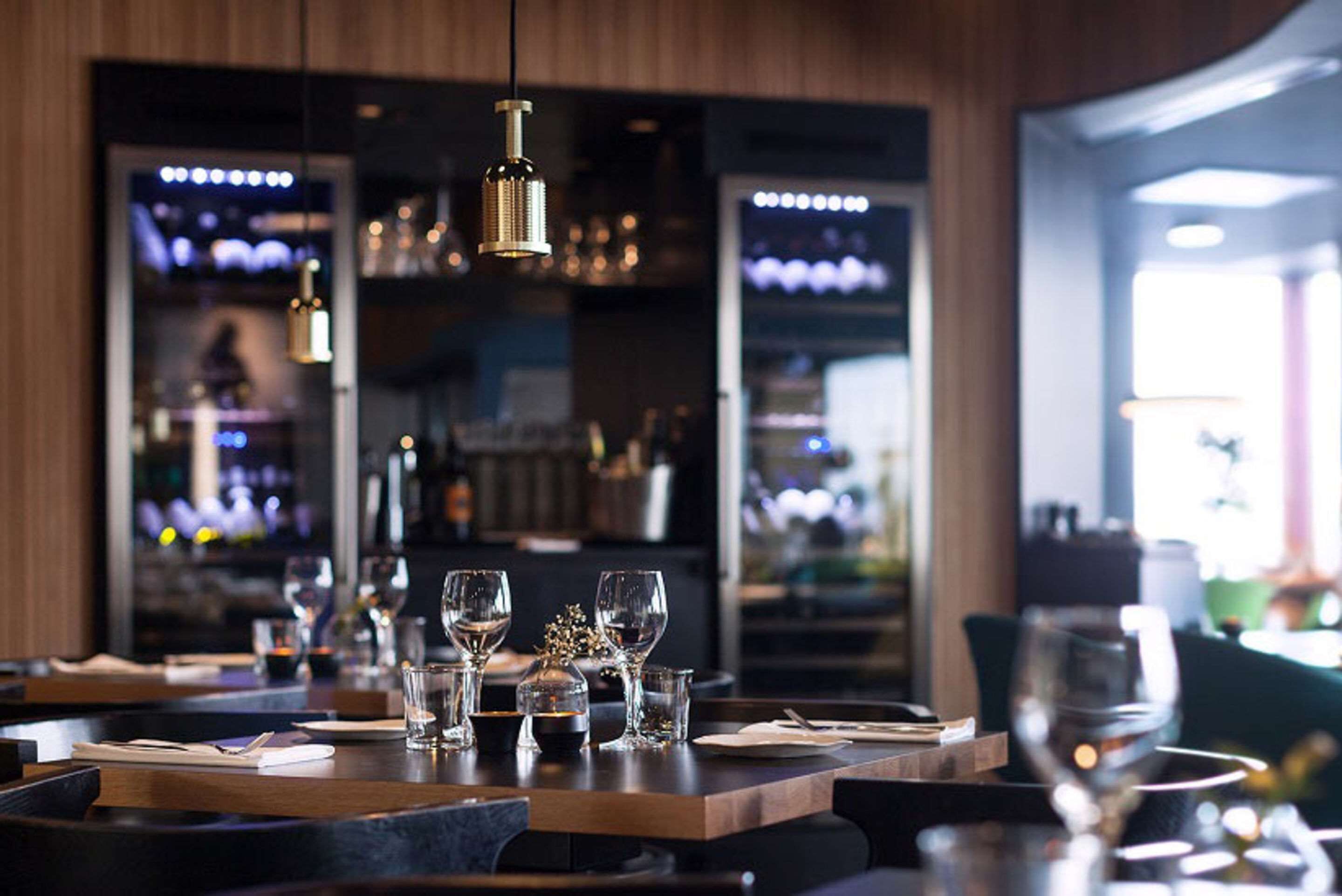 Interior de restaurante con mesas de madera, lámparas colgantes y una zona de bar al fondo.