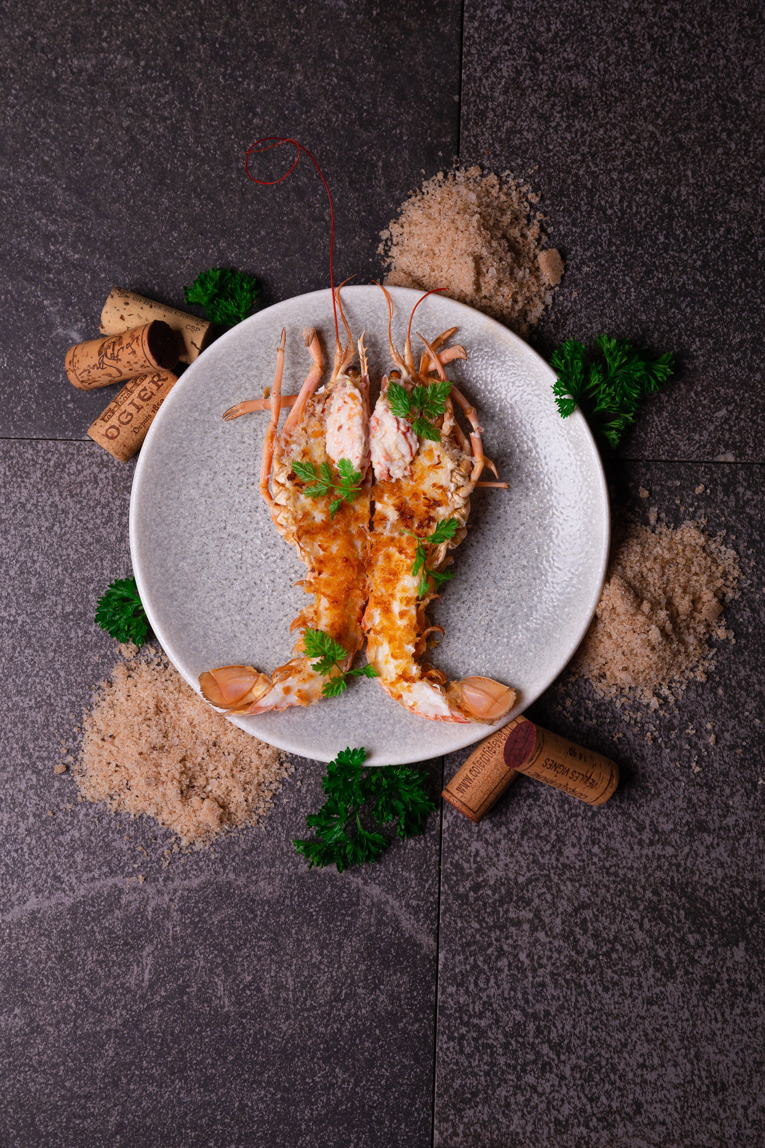 Une assiette sur laquelle repose un homard cuit, le tout présenté sur une assiette grise à texture, et entouré de bouchons de liège et de persil éparpillés sur une surface sombre.