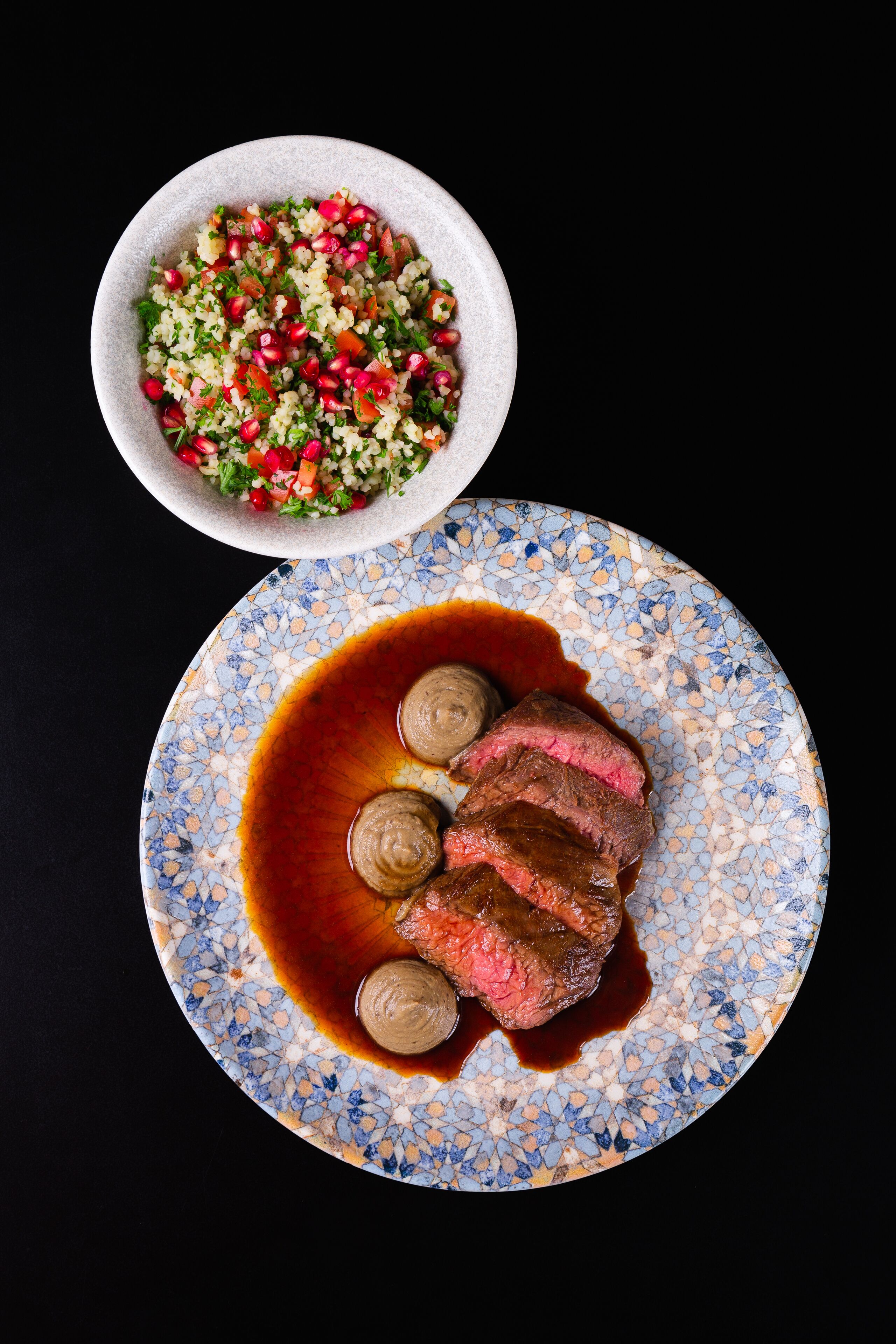 Une assiette de steak nappé d’une sauce brune et un saladier de salade parsemée de graines de grenade.