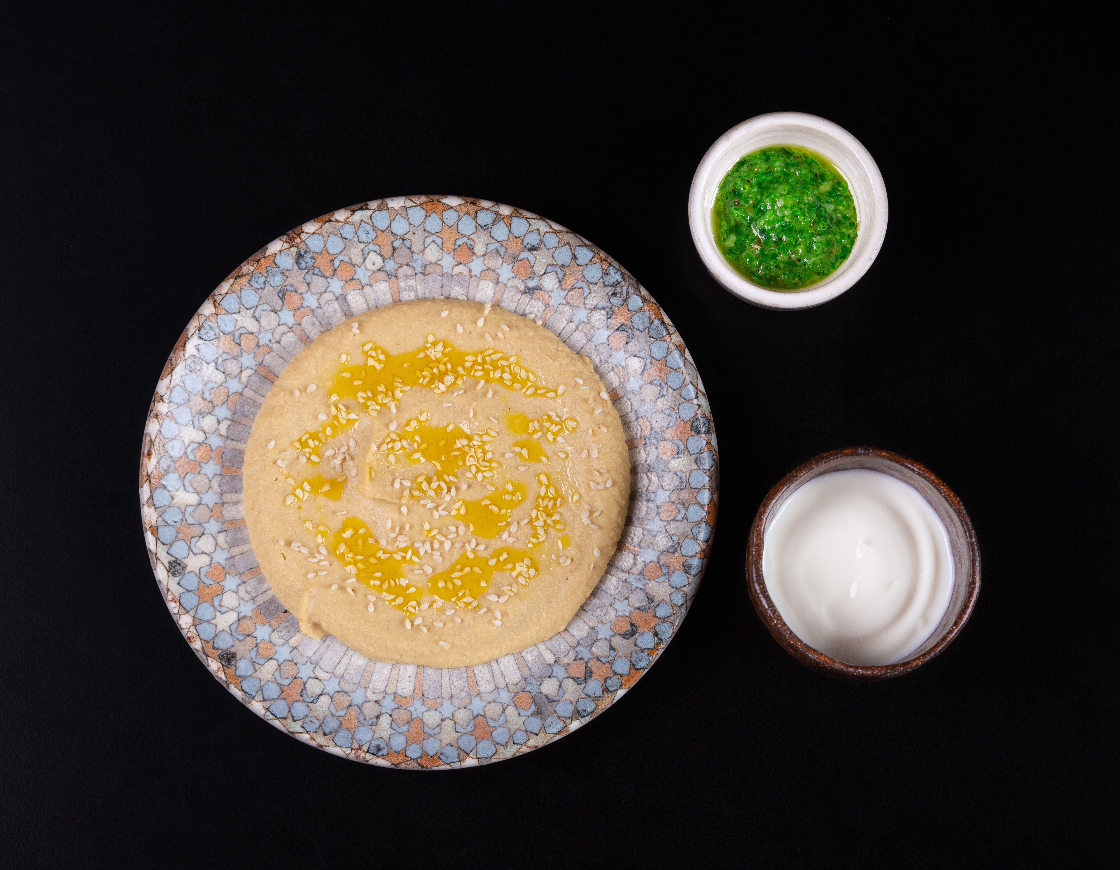 Une assiette de houmous parsemé de graines de sésame et arrosé d’huile d’olive, un petit bol de sauce verte et un verre de sauce blanche.