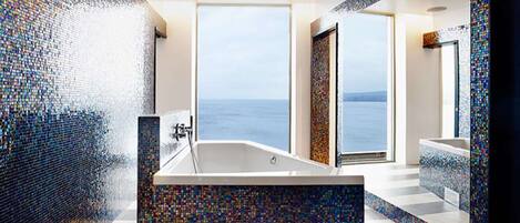 A modern bathroom with a mosaic-tiled wall, a large bathtub, and a view of the sea.