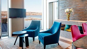 A modern living room with a large TV, two blue armchairs, a glass coffee table, and a view of a body of water. - Vox Hotel (Jonkoping)