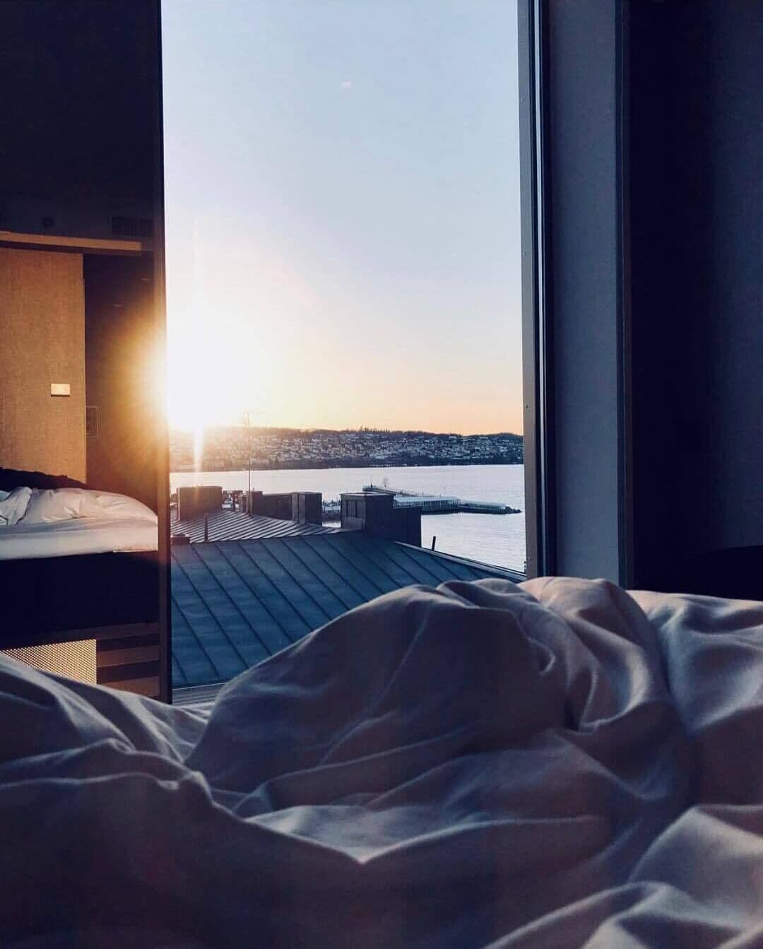 Una habitación de hotel con cama y un ventanal que da a una masa de agua y un horizonte urbano distante.