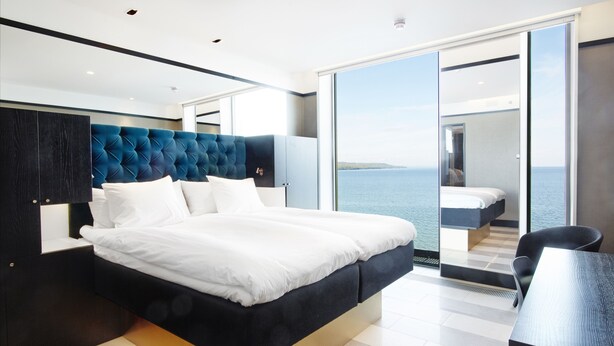 A modern bedroom with a large bed, a dark headboard, and a view of the sea through a sliding glass door. - Vox Hotel (Jonkoping)