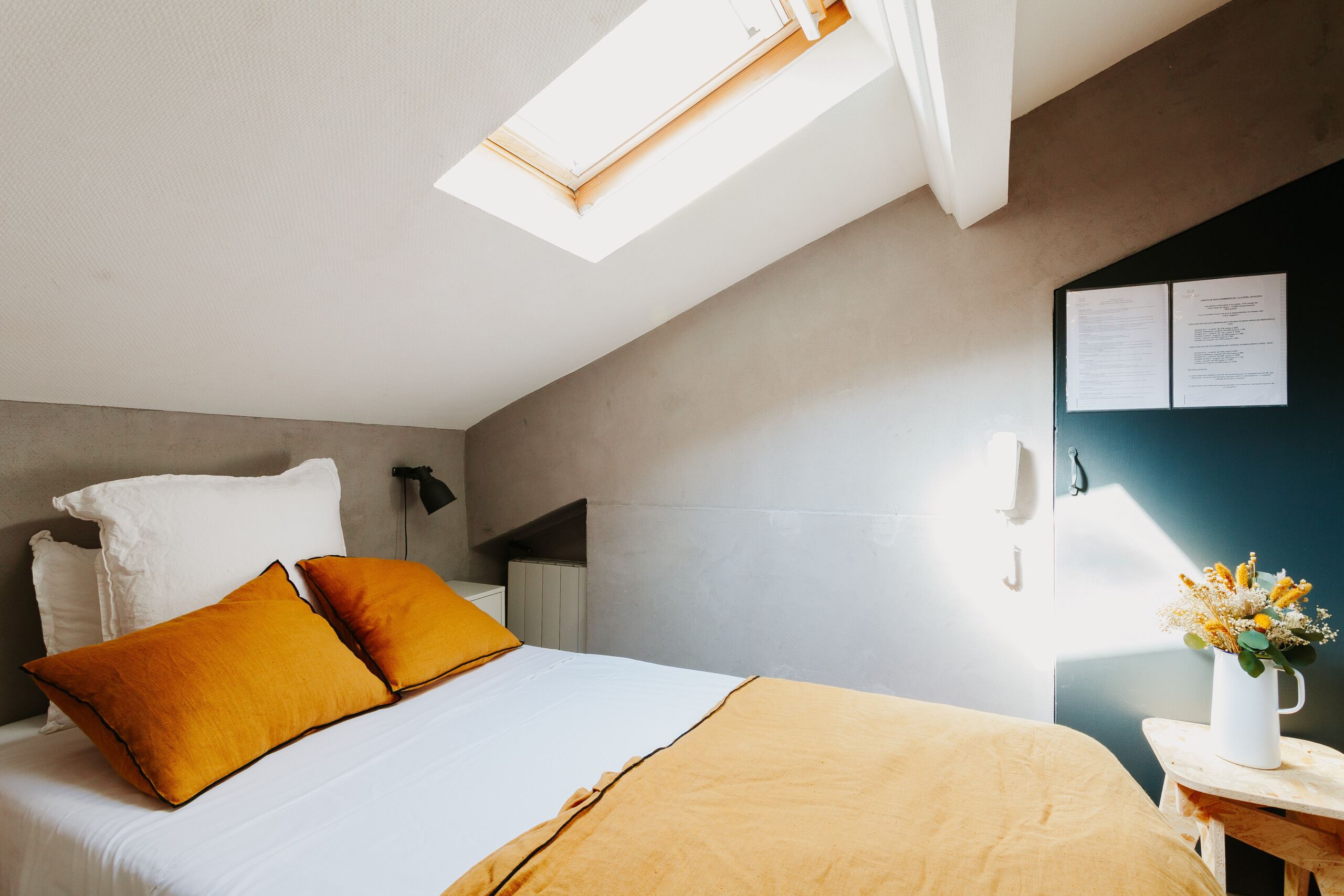 A modern hotel room with a skylight, a bed with white and orange bedding, a nightstand with a vase of flowers, and a wall-mounted phone.