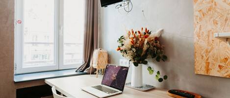 A hotel room with a wooden desk, a laptop, a remote control, a vase with flowers, a wall-mounted TV, and a window with curtains.