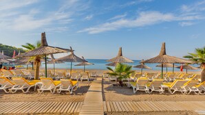 Een strand met rijen ligbedden en rieten parasols, een houten looppad en een helder uitzicht op de zee.