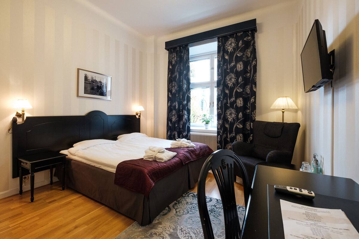 Habitación de hotel con cama, mesitas de noche, escritorio, silla, televisor y ventana con cortinas.