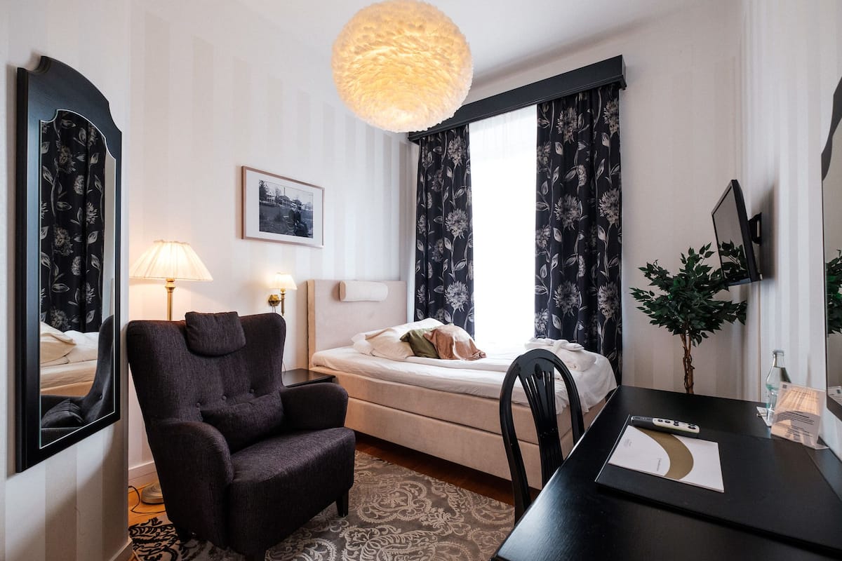 Habitación de hotel con cama, una silla, escritorio con control remoto, lámpara, planta y un espejo grande.
