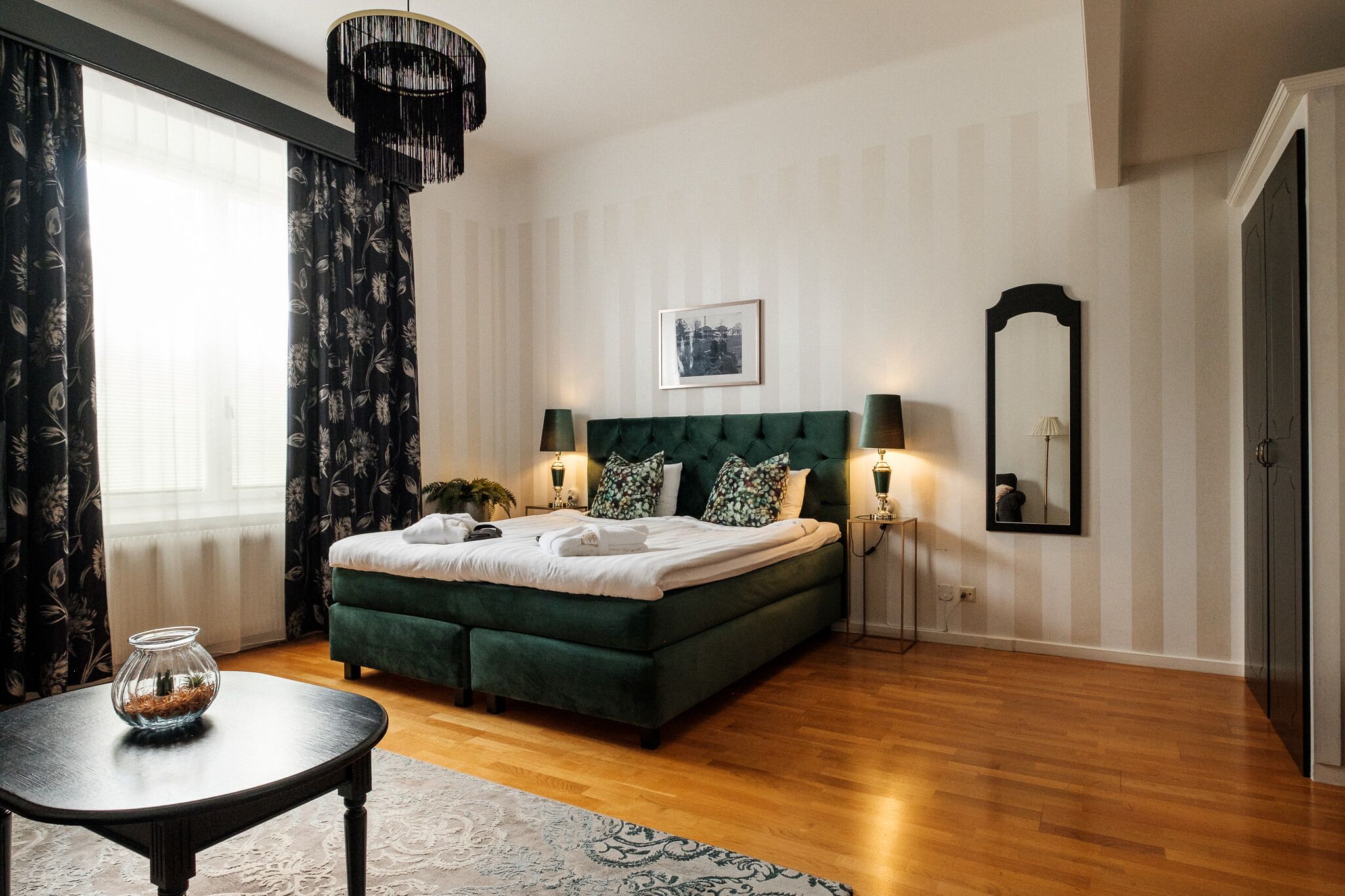 A bedroom with a green upholstered bed, two bedside lamps, a framed picture on the wall, a mirror, and a small table with a vase.