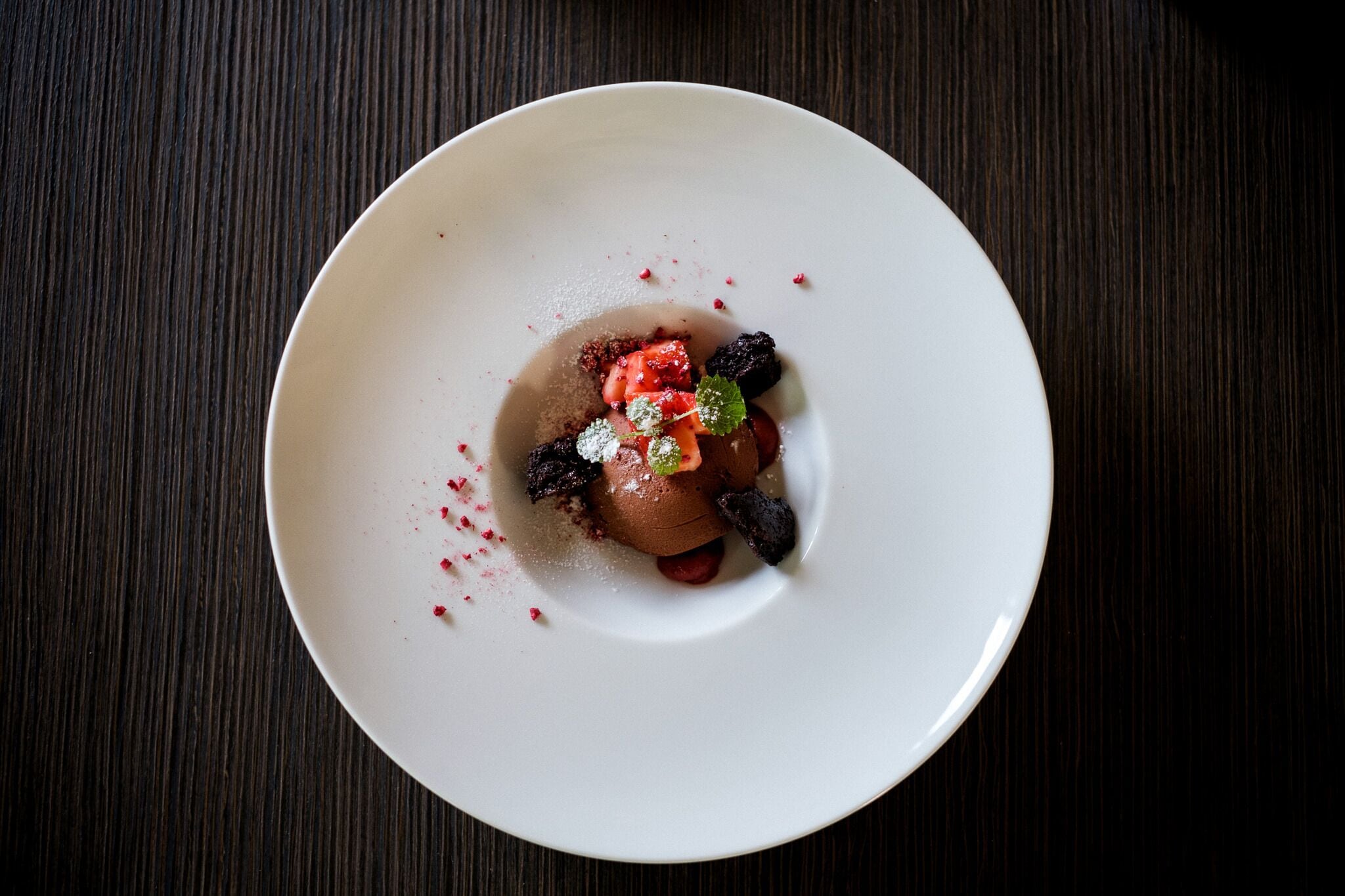 A dessert on a white plate with berries and mint on a dark wooden table.