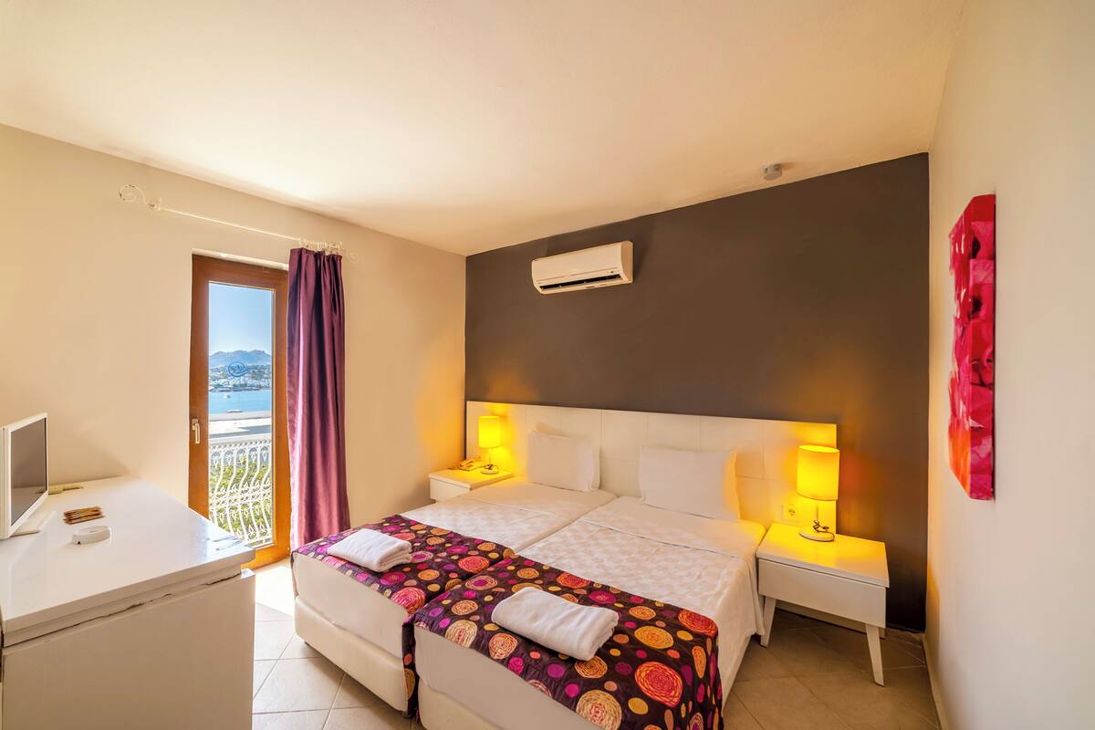 A hotel room with a bed, bedside lamps, a desk with a computer, and a view of the sea.