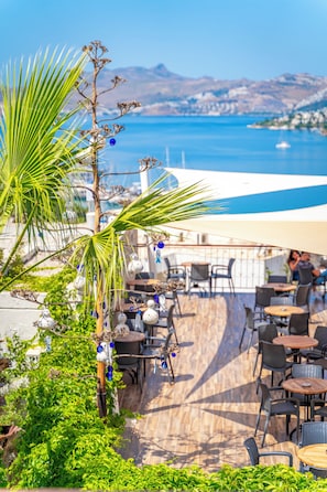 An outdoor seating area with tables and chairs, a palm tree, and a view of the sea and mountains. - Riva Bodrum Resort (Bodrum)