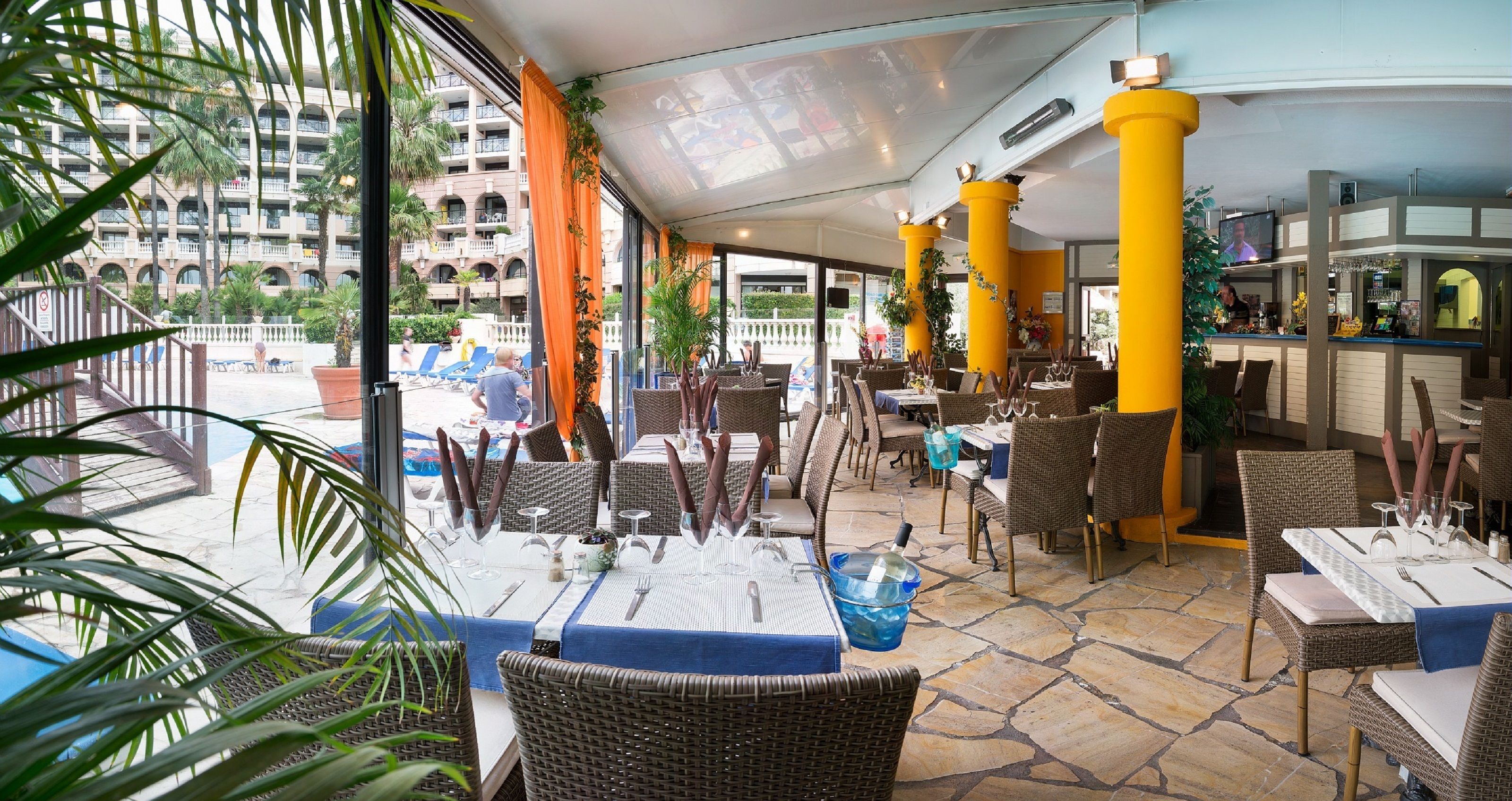 An outdoor dining area with wicker chairs and tables, yellow pillars, and a view of a hotel building.