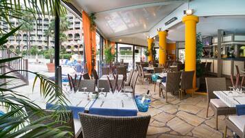 An outdoor dining area with wicker chairs and tables, yellow pillars, and a view of a hotel building.