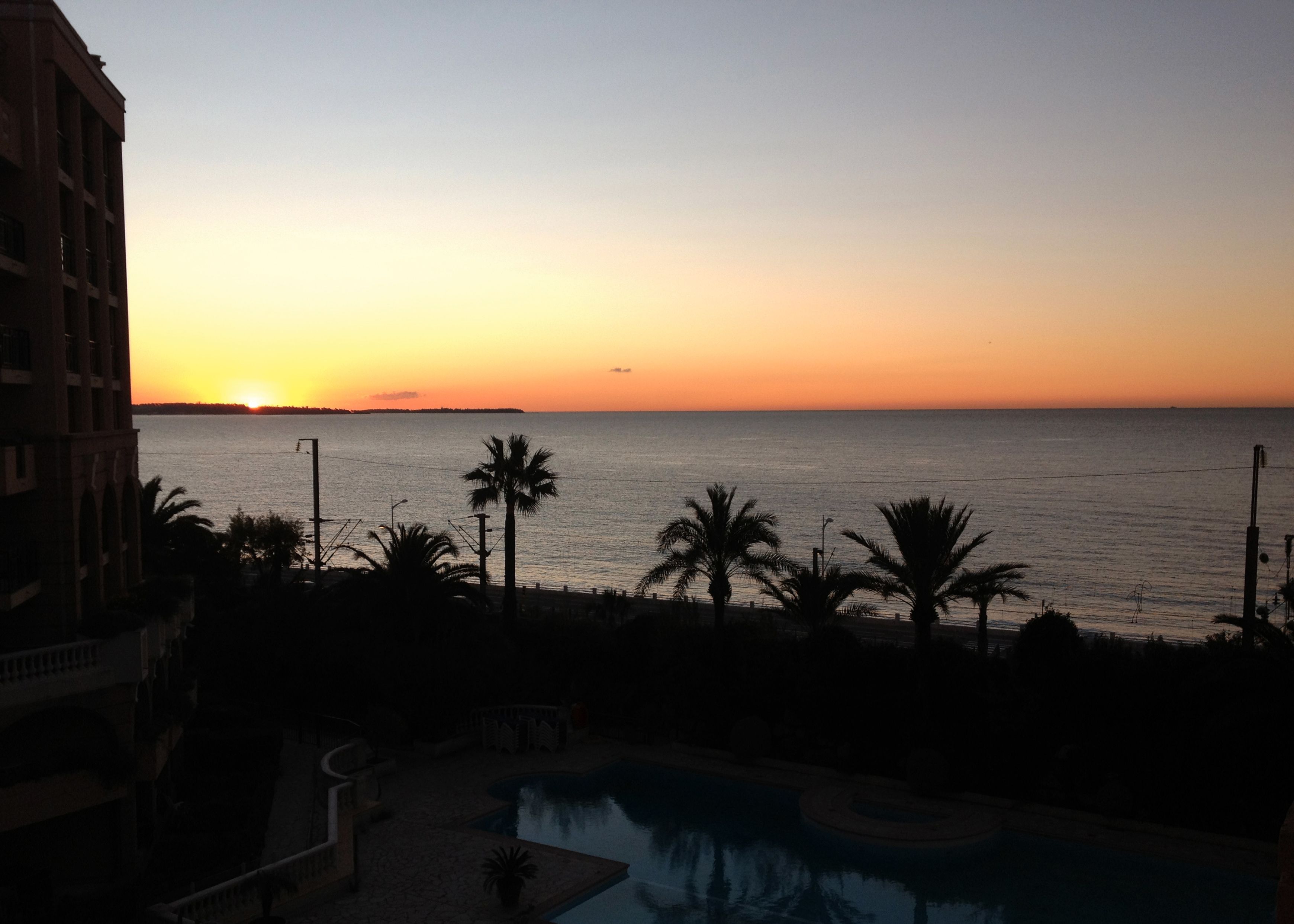 A resort with a pool, palm trees, and a view of the sunset over the sea.