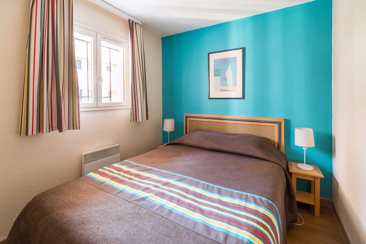 Un dormitorio con cama, ventana con cortinas, mesita de noche con lámpara y un cuadro en la pared.