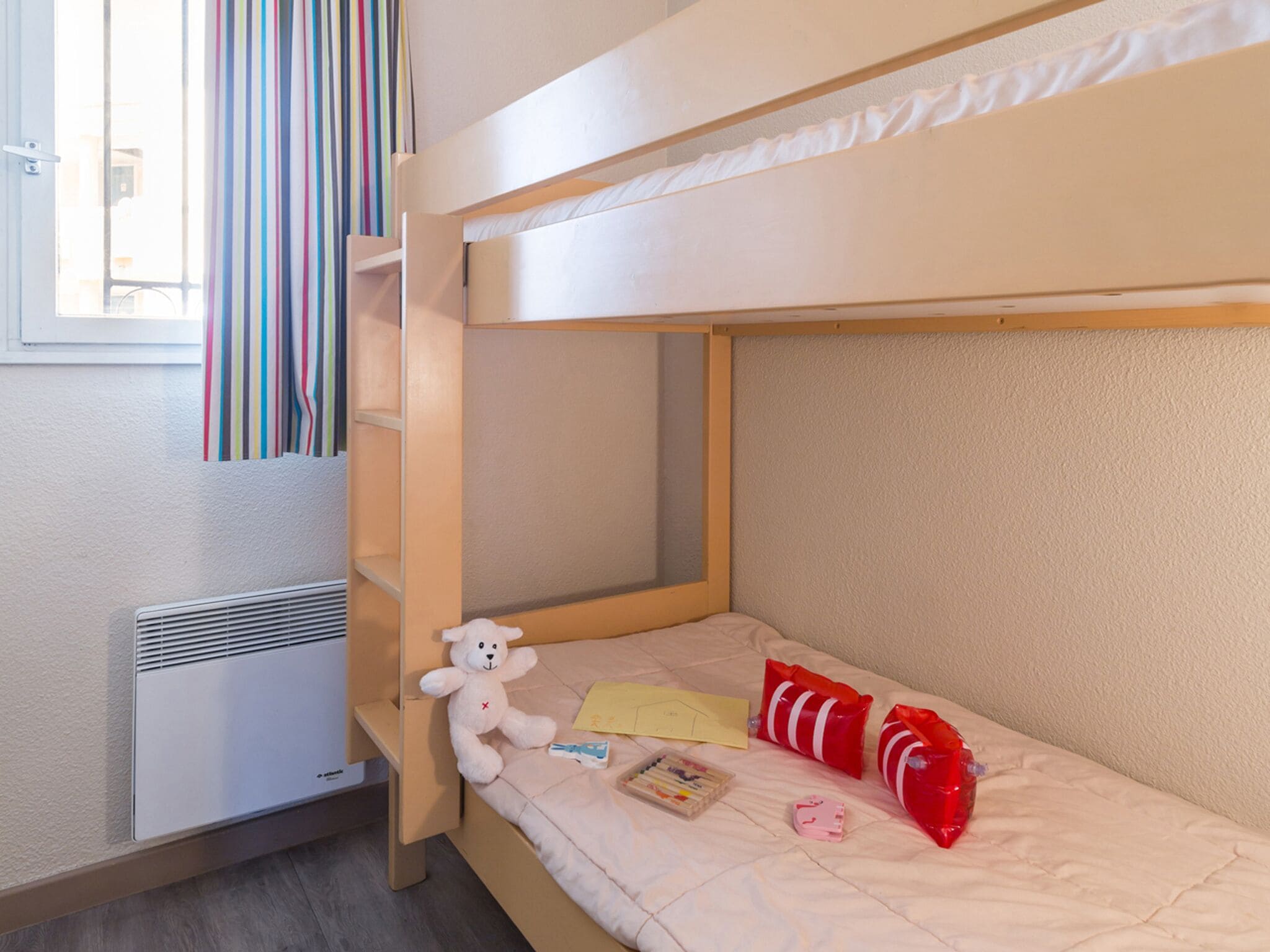Habitación con literas, ventana, cortinas a rayas, un calentador, un oso de peluche y algunos objetos sobre la cama.