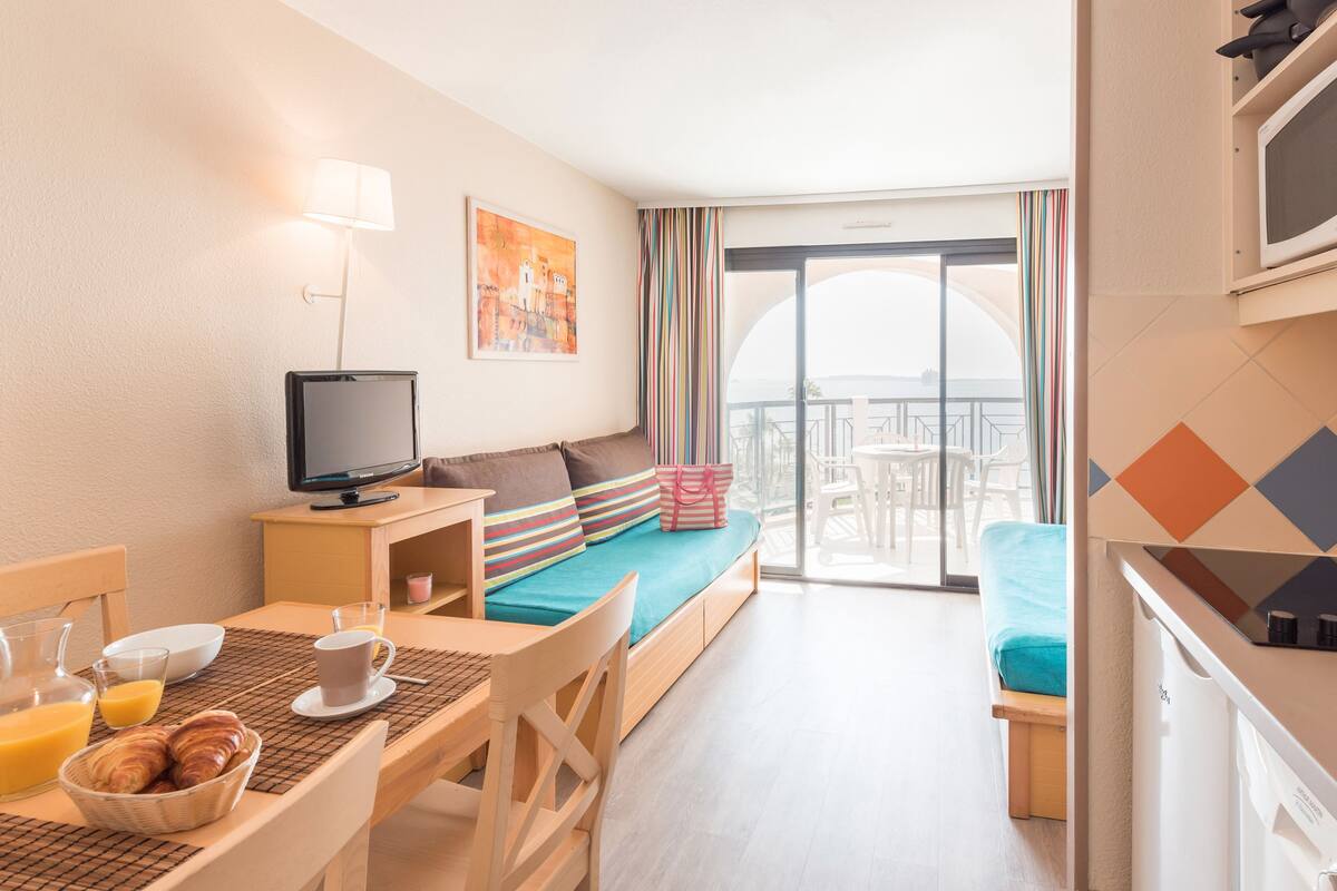 Habitación de hotel con cama, televisor, mesa de desayuno y balcón con vistas.