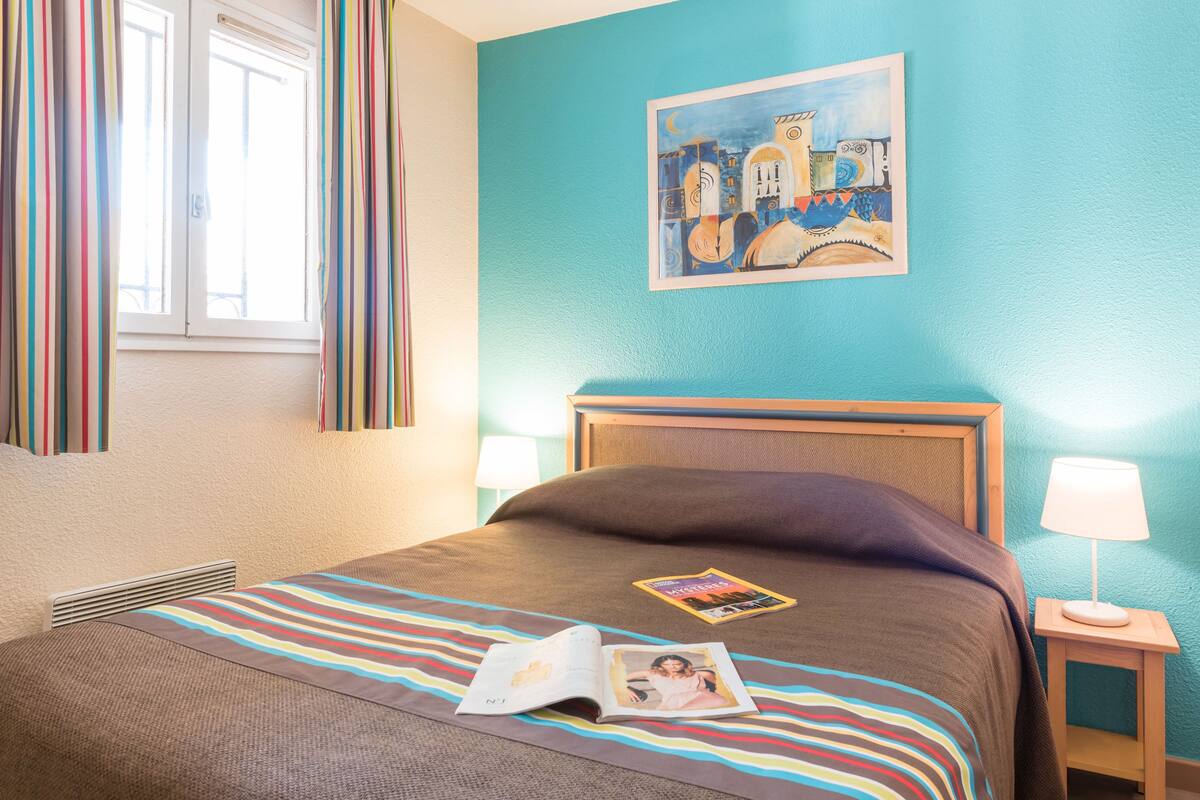 Habitación de hotel con cama, ventana con cortinas, un cuadro en la pared, lámpara de noche y una revista sobre la cama.