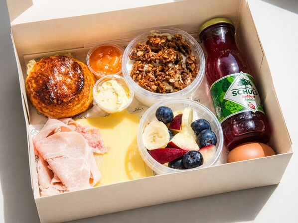 Une boîte de petit-déjeuner contenant un croissant, un yaourt, du granola, des fruits, du fromage, du jambon et une bouteille de jus de fruits.