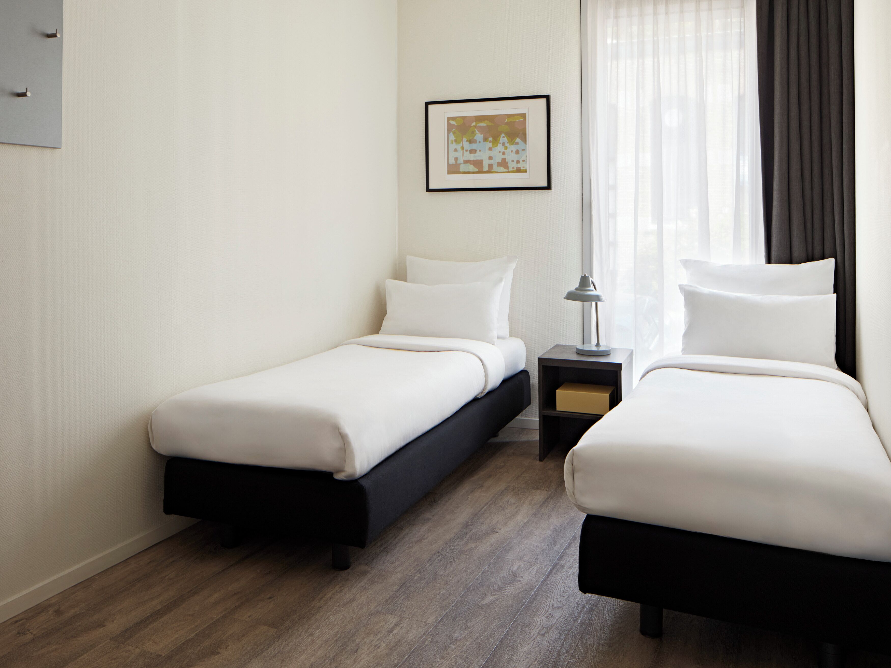 A hotel room with two single beds, a nightstand with a lamp, a framed picture on the wall, and a window with curtains.