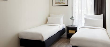 A hotel room with two single beds, a nightstand with a lamp, a framed picture on the wall, and a window with curtains.