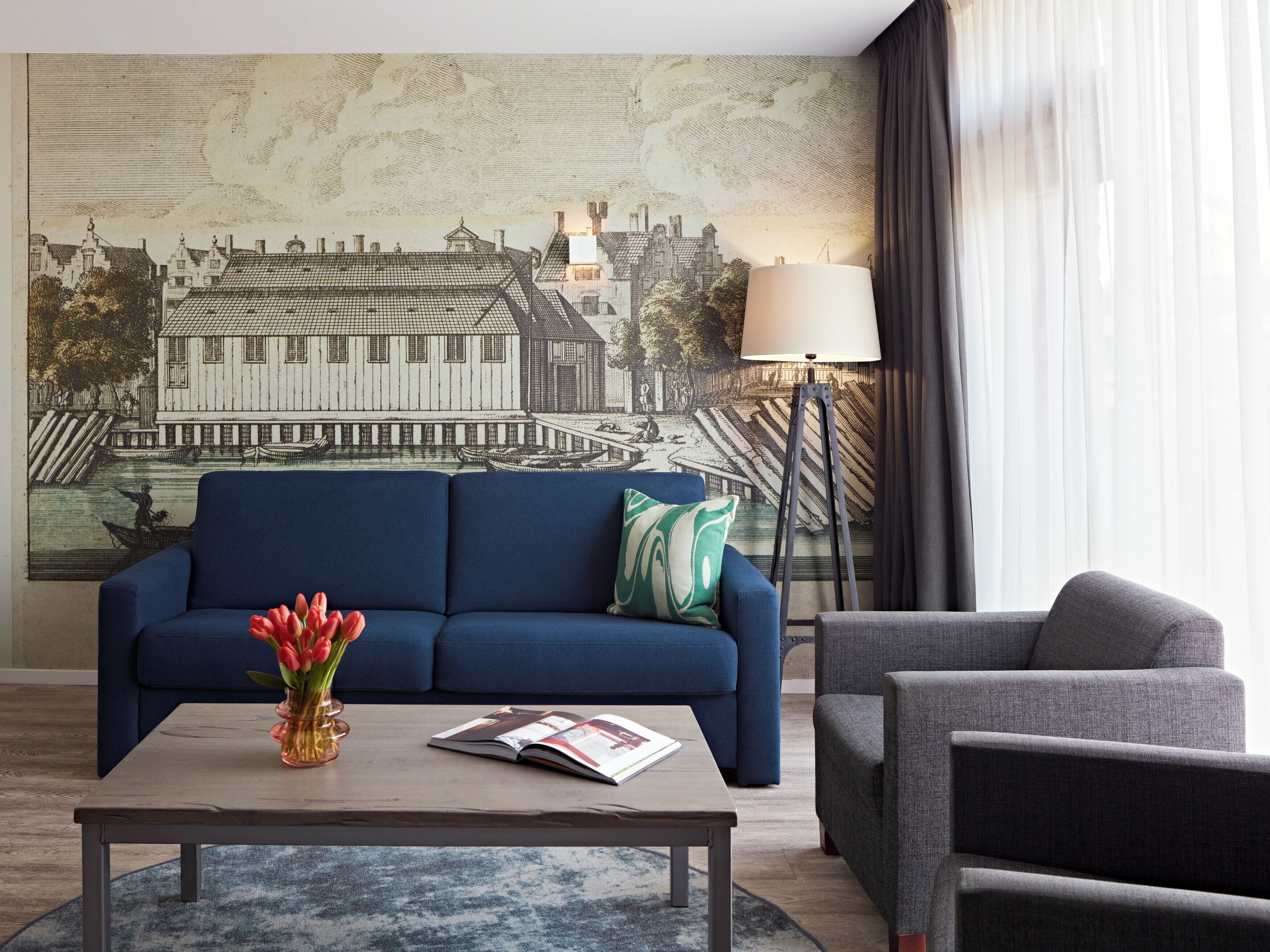 A living room with a blue sofa, a grey armchair, a coffee table with a book and a vase of red tulips, a floor lamp, and a large cityscape mural on the wall.