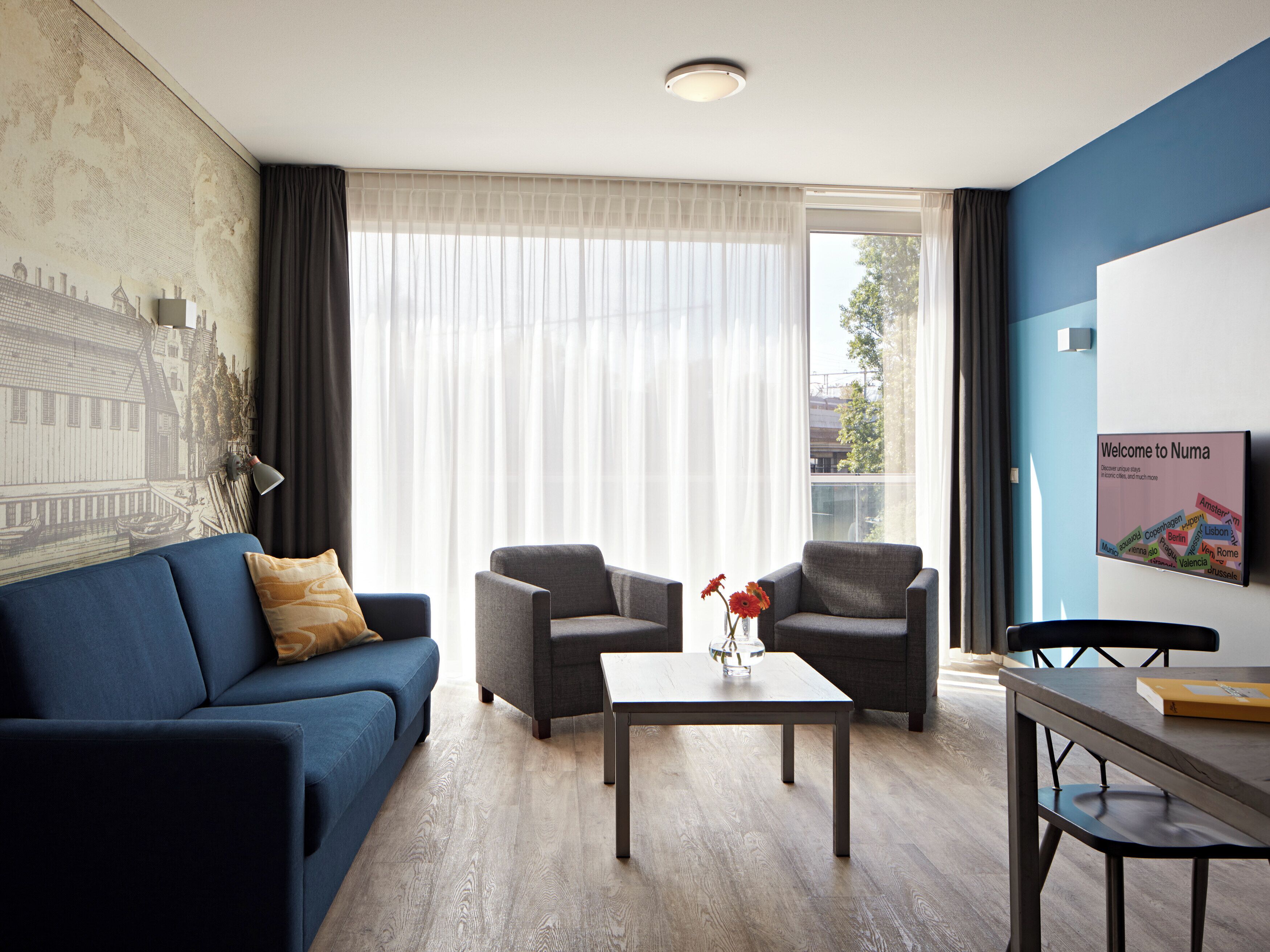 Una sala moderna con un sofá azul, dos sillones, una mesa de centro y un mural con vistas a la ciudad en la pared.