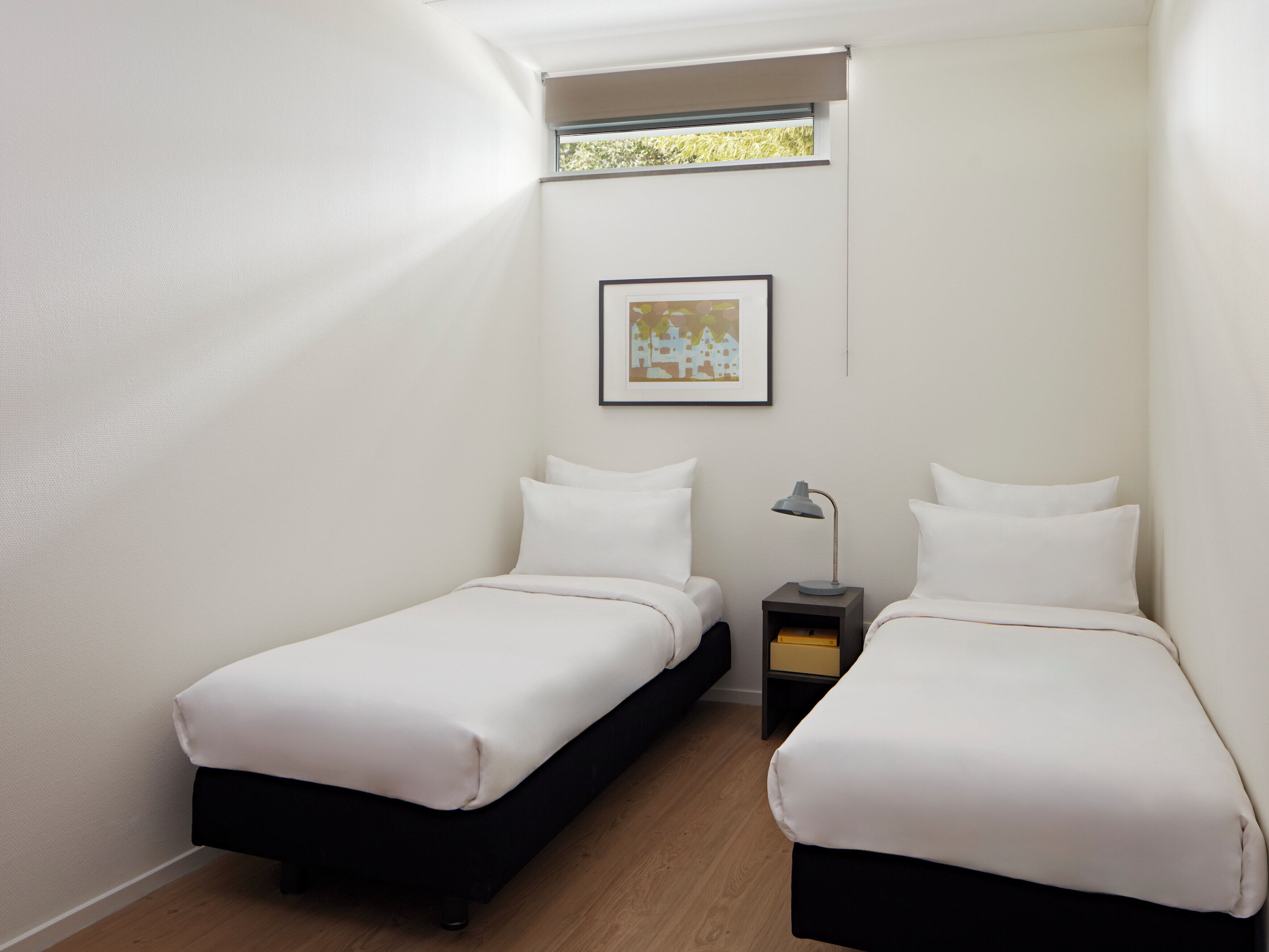 Una habitación pequeña y moderna con dos camas individuales, una mesita de noche con lámpara, un cuadro en la pared y una ventana con vistas a zonas verdes.