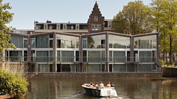 A modern building with large windows and a boat docked in front. - YAYS Amsterdam Prince Island by Numa (Amsterdam)