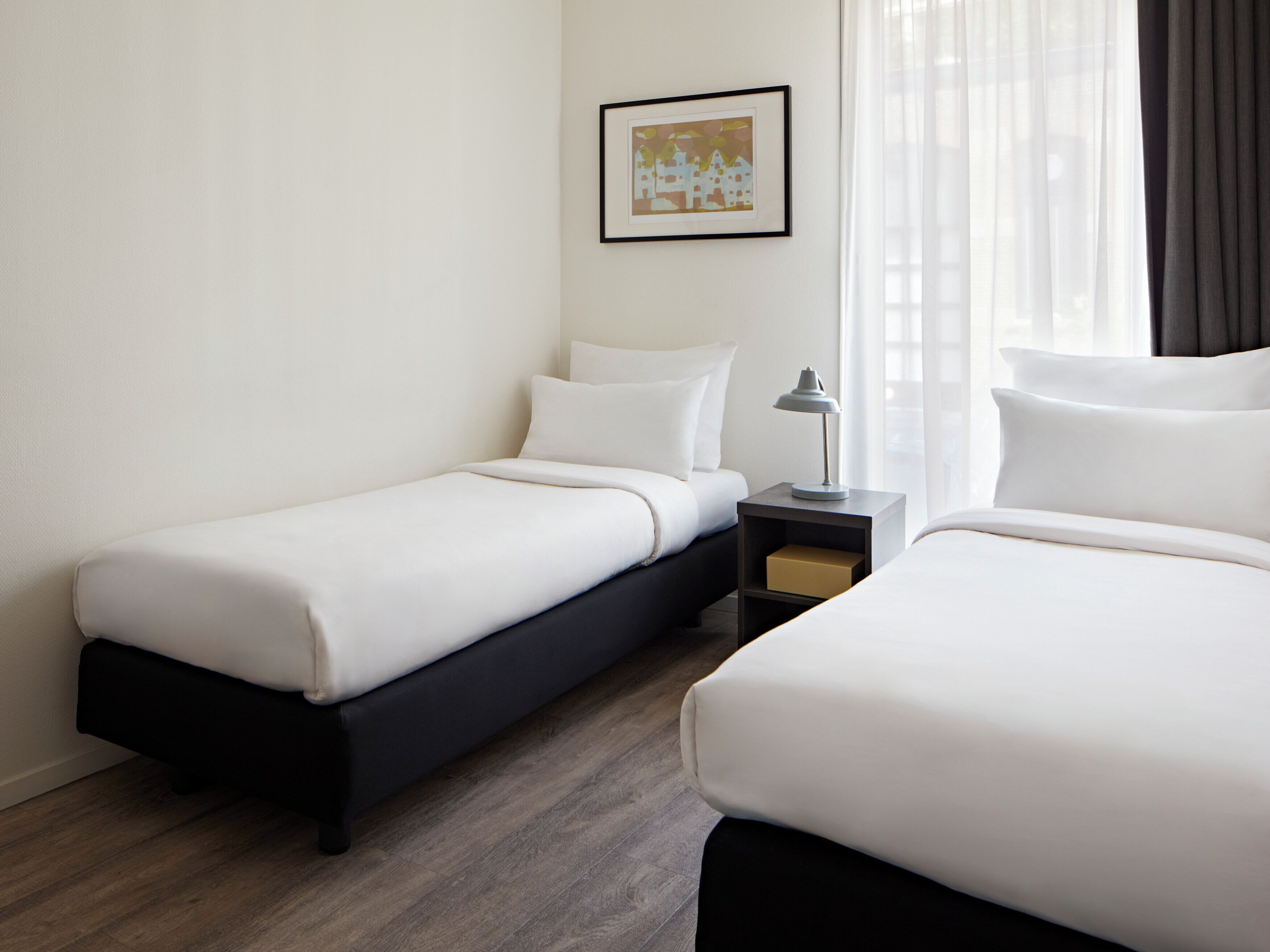 A hotel room with two single beds, a nightstand with a lamp, a framed picture on the wall, and a window with curtains.