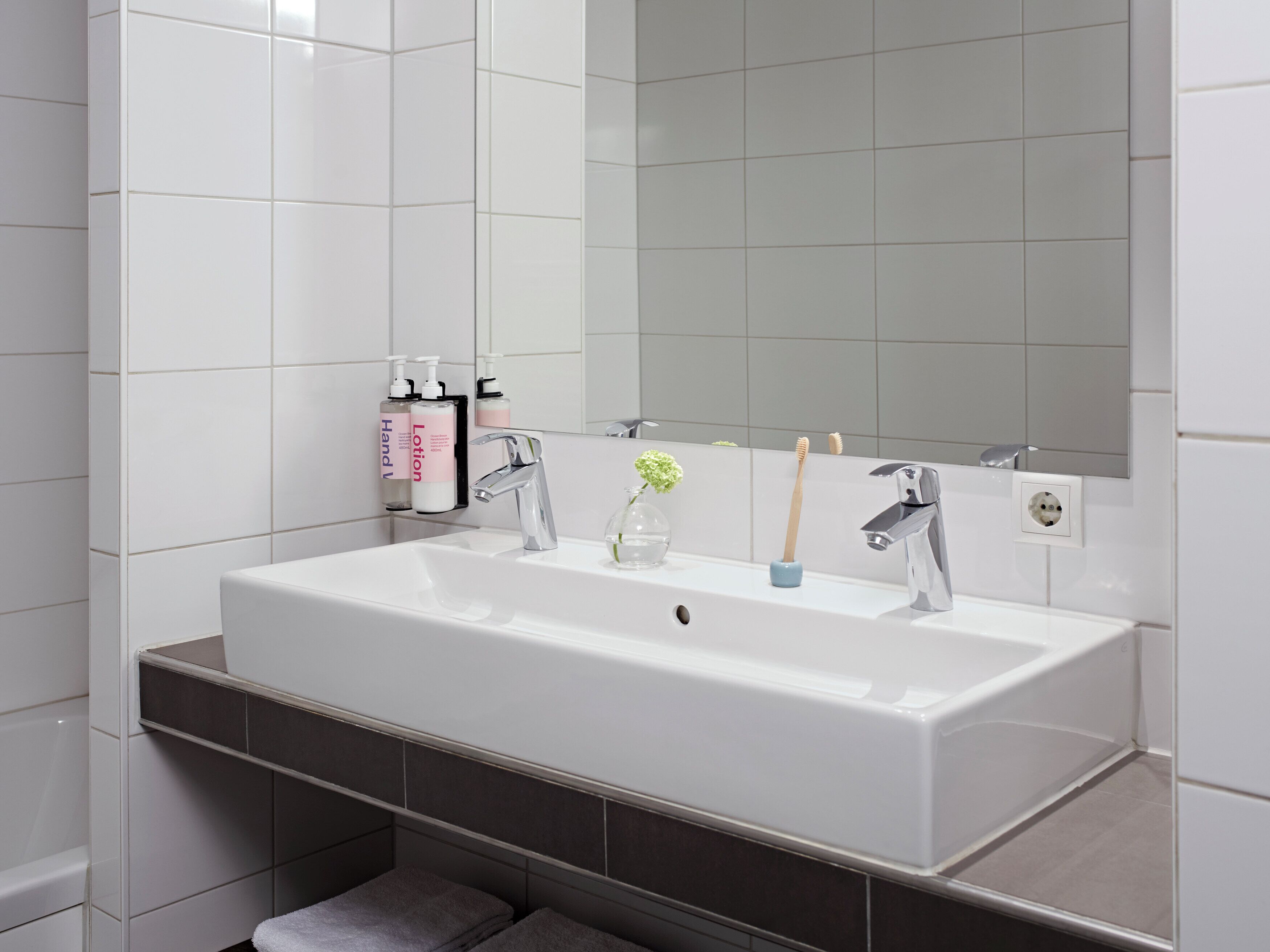 Ein Badezimmer mit einem weißen Waschbecken, zwei Wasserhähnen, einem Spiegel und Pflegeprodukten auf einem Regal.