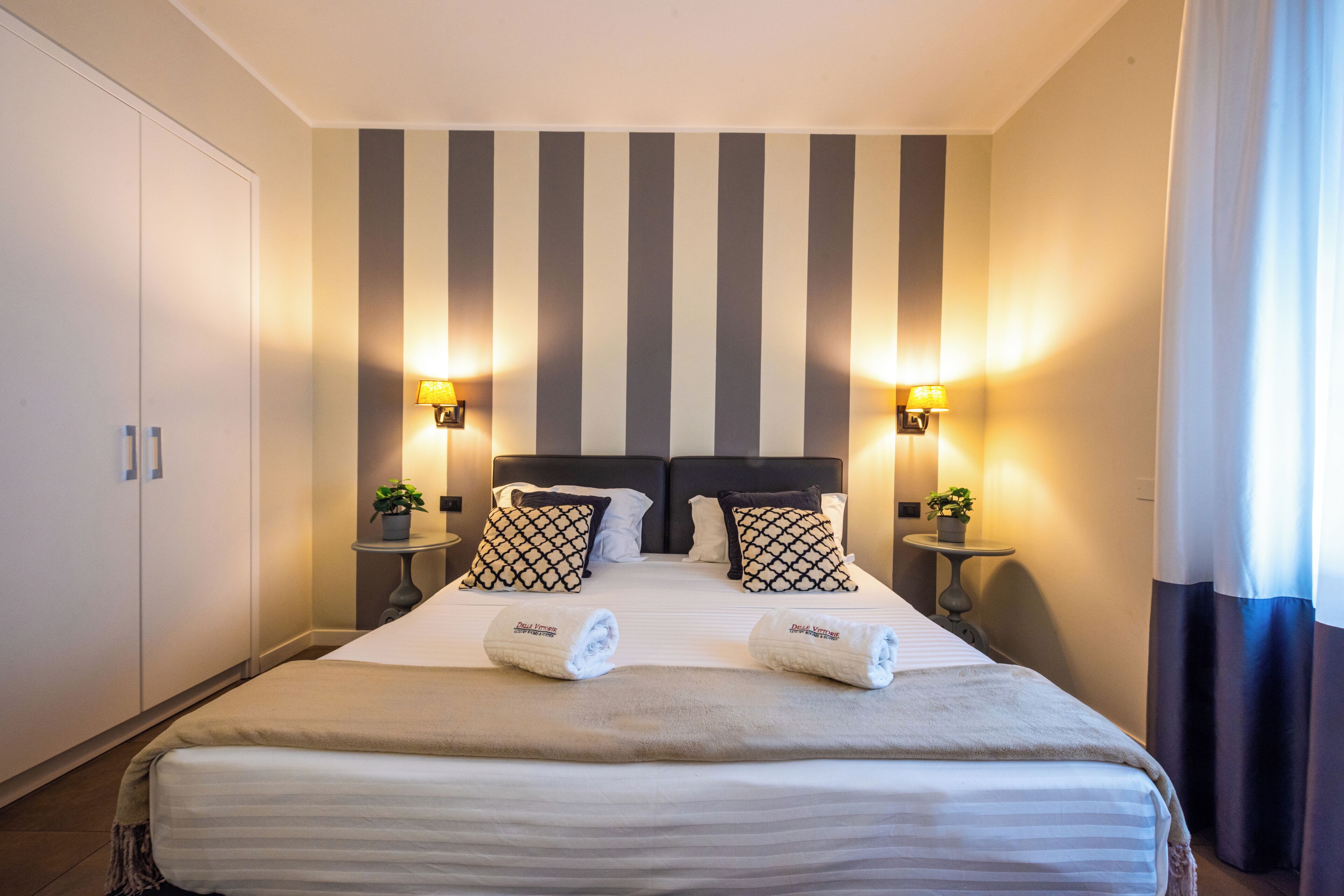 A hotel room with a large bed, striped wall, two bedside tables with lamps, and a wardrobe.