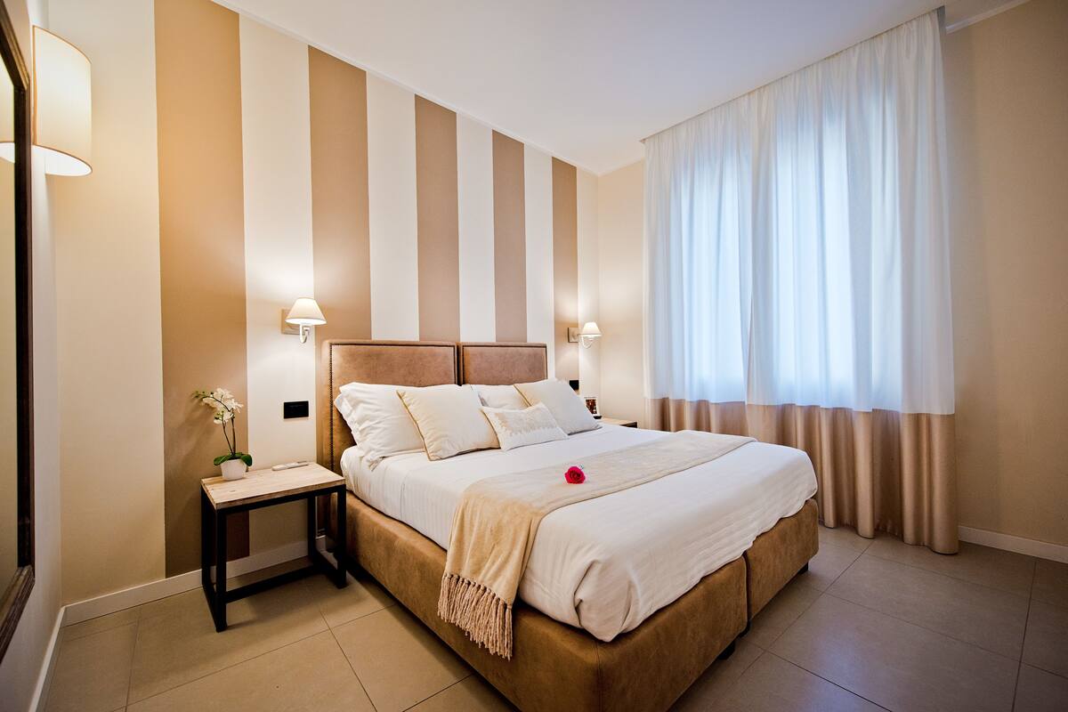 Une chambre d’hôtel avec un grand lit, deux tables de chevet, une petite table, une plante en pot et une fenêtre avec des rideaux.