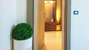 Ein Hotel mit Eingangsbereich, einer großen Pflanze in einem Topf, einer Holztür und einem Flur zu einem Zimmer mit Bett und Nachttisch.