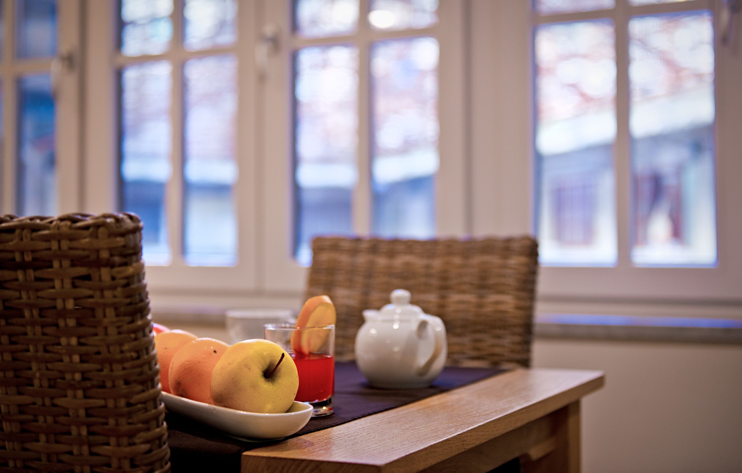 A wicker basket, a plate with fruits, a teapot, and a glass of red drink on a table by a window.