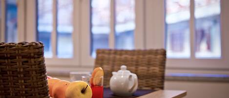 A wicker basket, a plate with fruits, a teapot, and a glass of red drink on a table by a window.