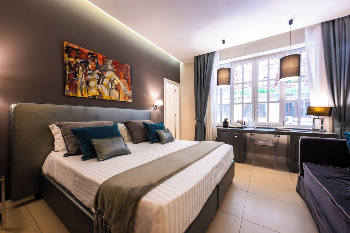 Une chambre moderne avec un grand lit, un bureau et une chaise. Il y a un tableau au mur et une fenêtre avec des rideaux.